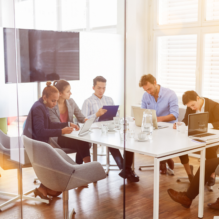 A diverse group of five people seated around a white conference table in a bright, modern office, working on laptops and tablets.