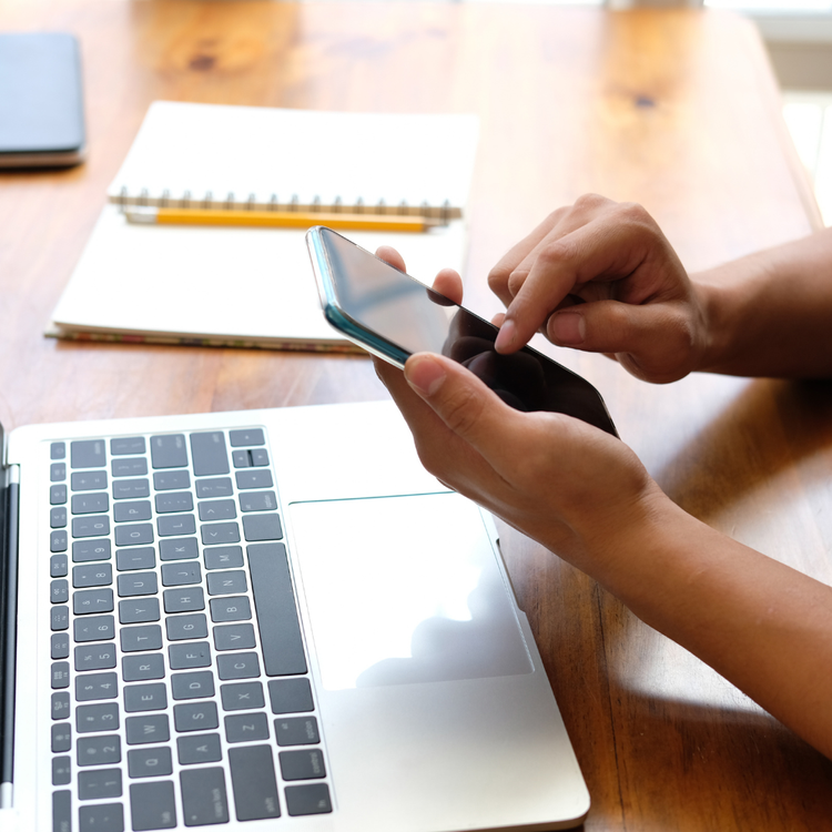 Person using a smartphone on a wooden desk with a laptop, notebook, and pen.
